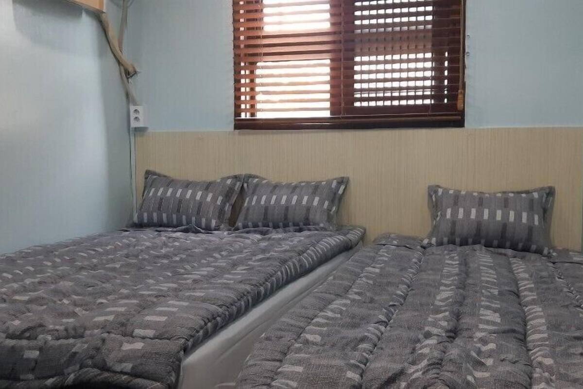 Un lit simple avec une couvre-lit à motifs et deux oreillers, une fenêtre avec des stores et des murs bleu clair.