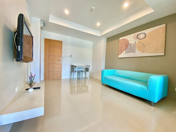 A modern living room with a teal sofa, a wooden door, a dining area, and a mounted flat-screen TV.