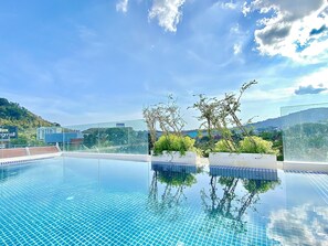 A clear blue swimming pool with a tiled floor, surrounded by greenery and a view of a cityscape with buildings and hills in the background.