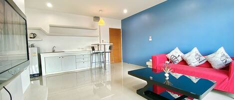 A modern living room with a sofa, a coffee table, and a TV. There is a kitchen area with a refrigerator and a kettle.