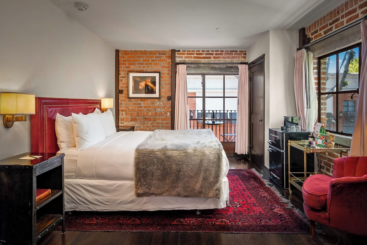 A hotel room with a large bed, a red headboard, a red rug, a small table with a lamp, and a brick wall.