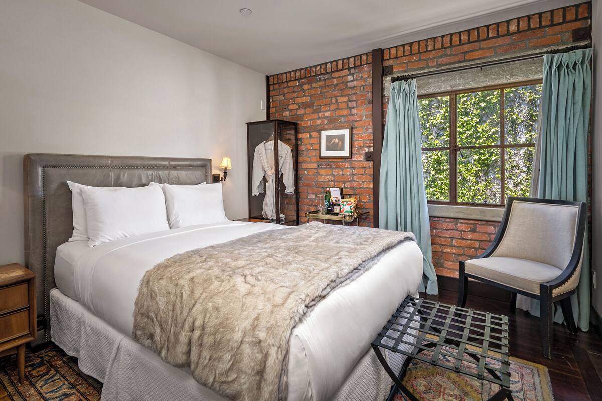 A hotel room with a large bed, a chair, a brick wall, and a window with curtains.