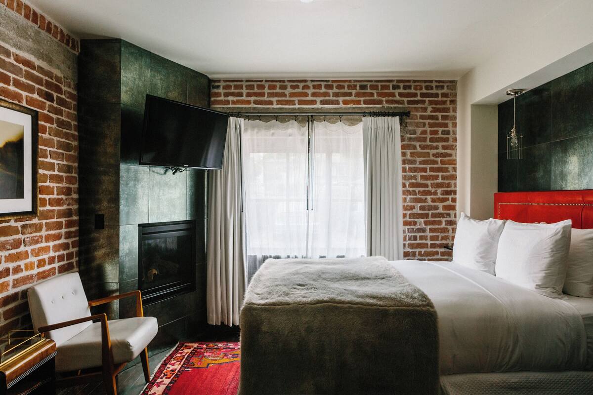 A hotel room with a brick wall, a flat-screen TV, a white bed, a red headboard, and a wooden chair.