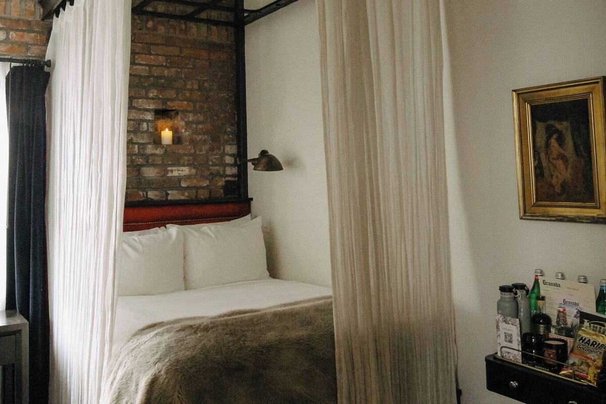 A four-poster bed with white linens, a brick wall, a small table with bottles, and a framed picture on the wall.