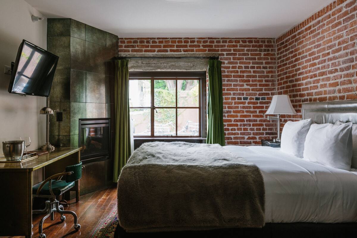 A hotel room with a brick wall, a bed with white linens, a desk with a chair, and a television mounted on the wall.