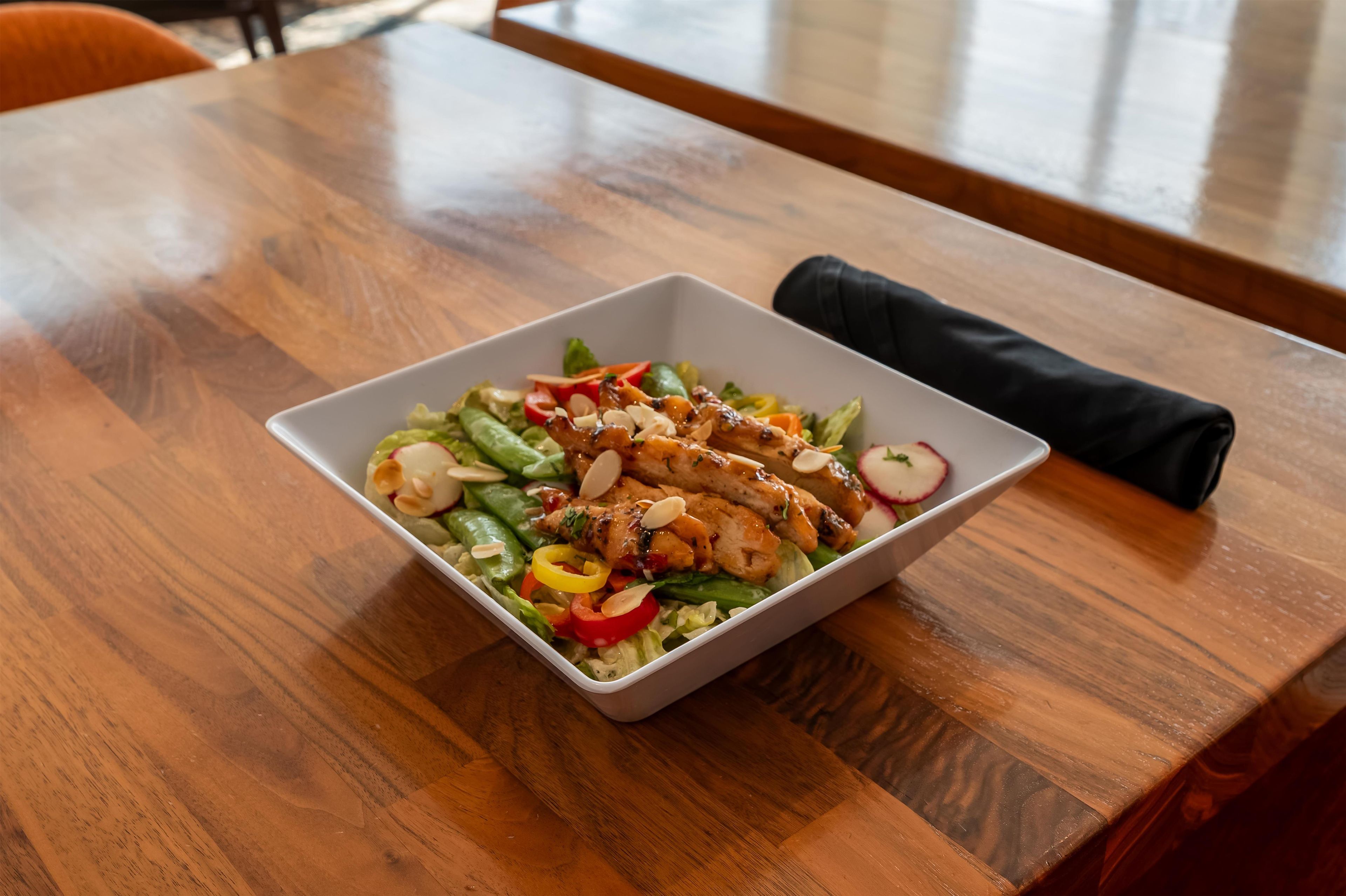 A square white bowl on a wooden table with a black napkin beside it, containing a salad with greens, red peppers, and sliced almonds.