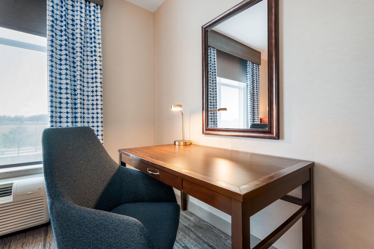 Quarto de hotel com escrivaninha de madeira, cadeira, abajur, espelho grande e janela com cortinas estampadas.