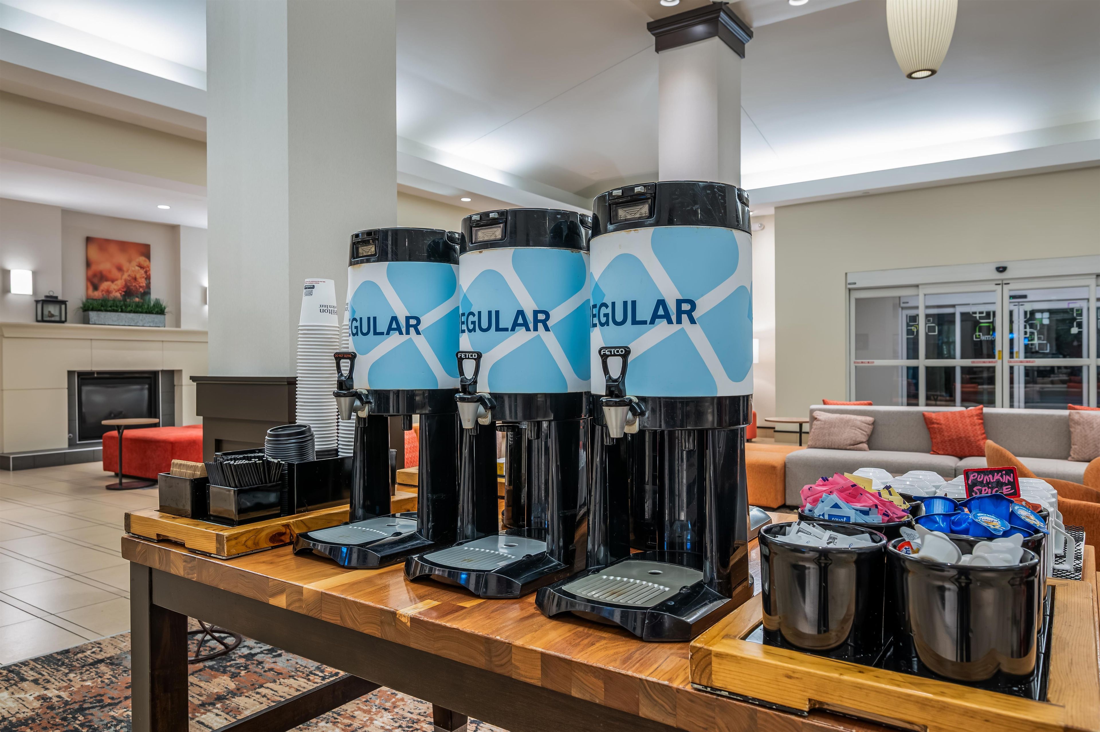 Una estación de café con varios dispensadores de café etiquetados como 'Regular', en un moderno vestíbulo con un sofá y una chimenea al fondo.