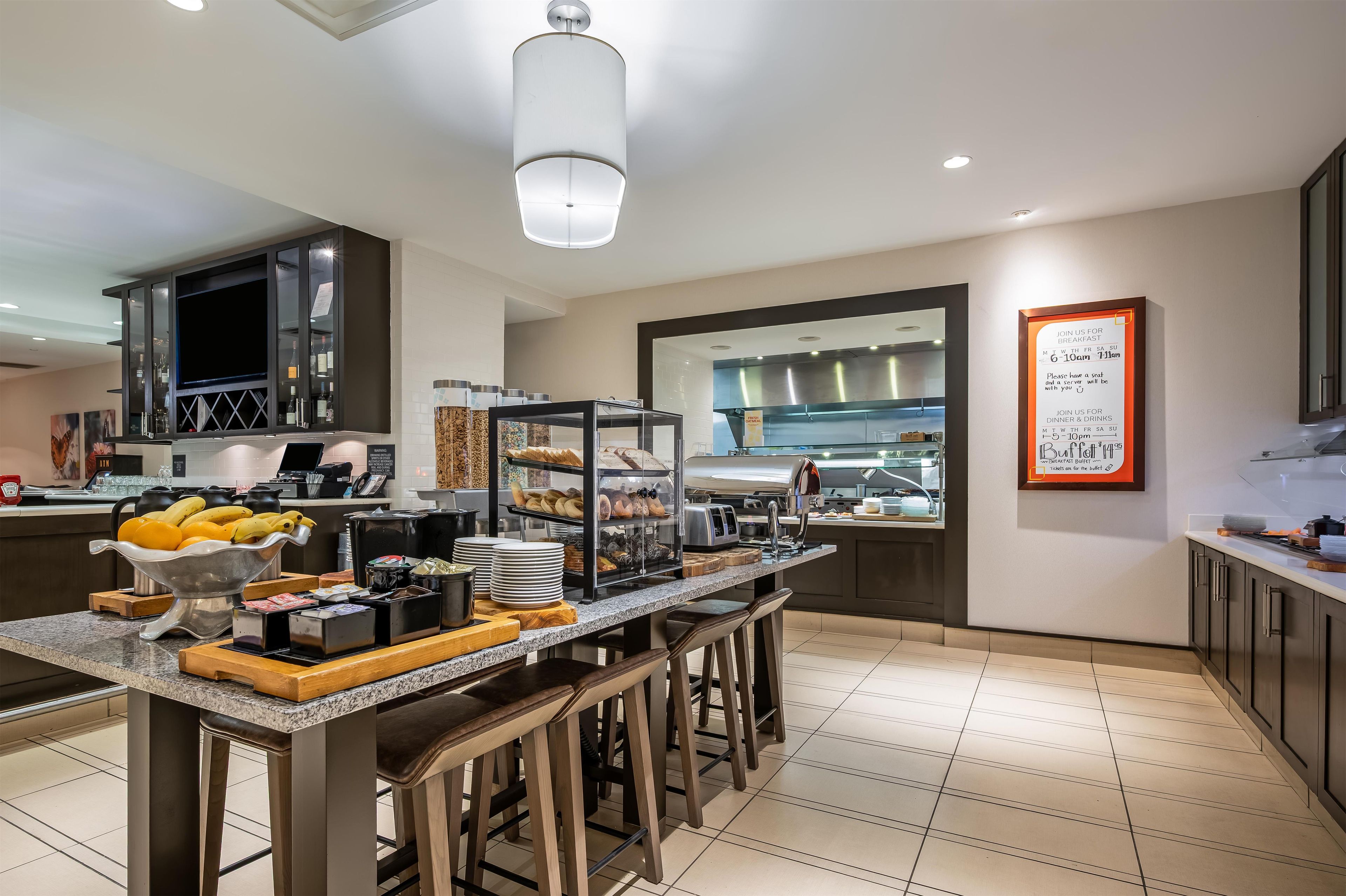 A modern breakfast buffet with a variety of food items, a seating area with stools, and a clear view of the kitchen in the background.