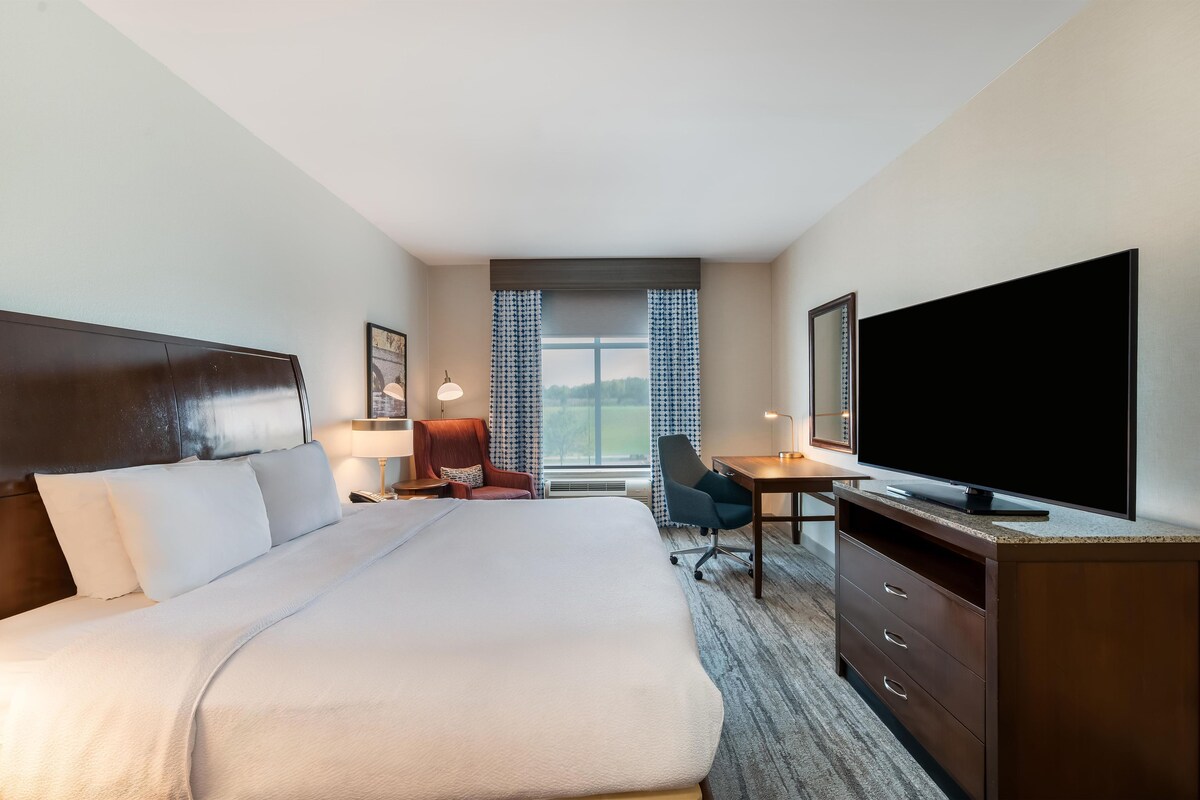 Quarto de hotel com uma cama grande, uma mesa com televisão de tela plana, uma cadeira e uma janela com cortinas.