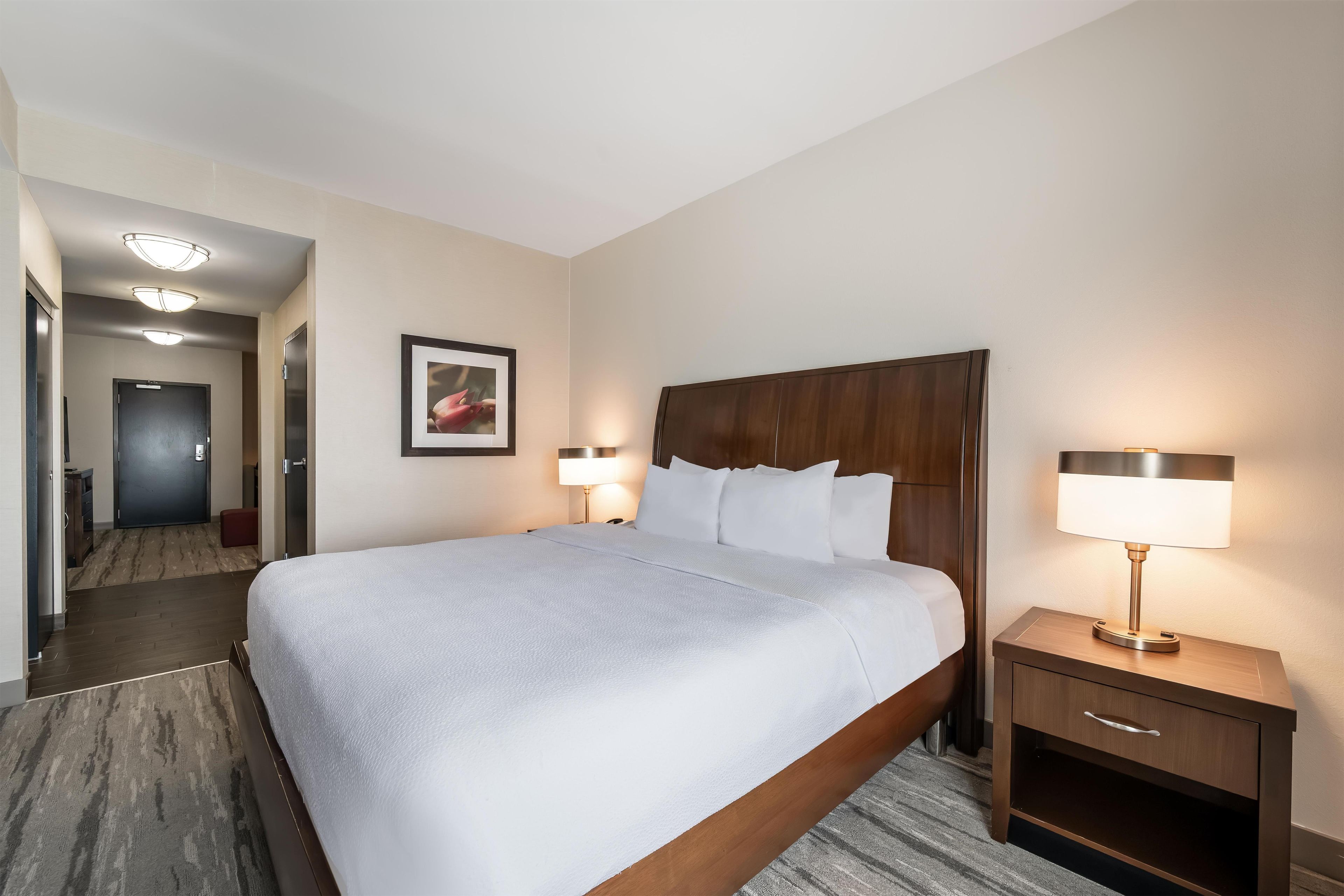 A hotel room with a large bed, two bedside lamps, a wooden headboard, a framed picture on the wall, and a door leading to another room.