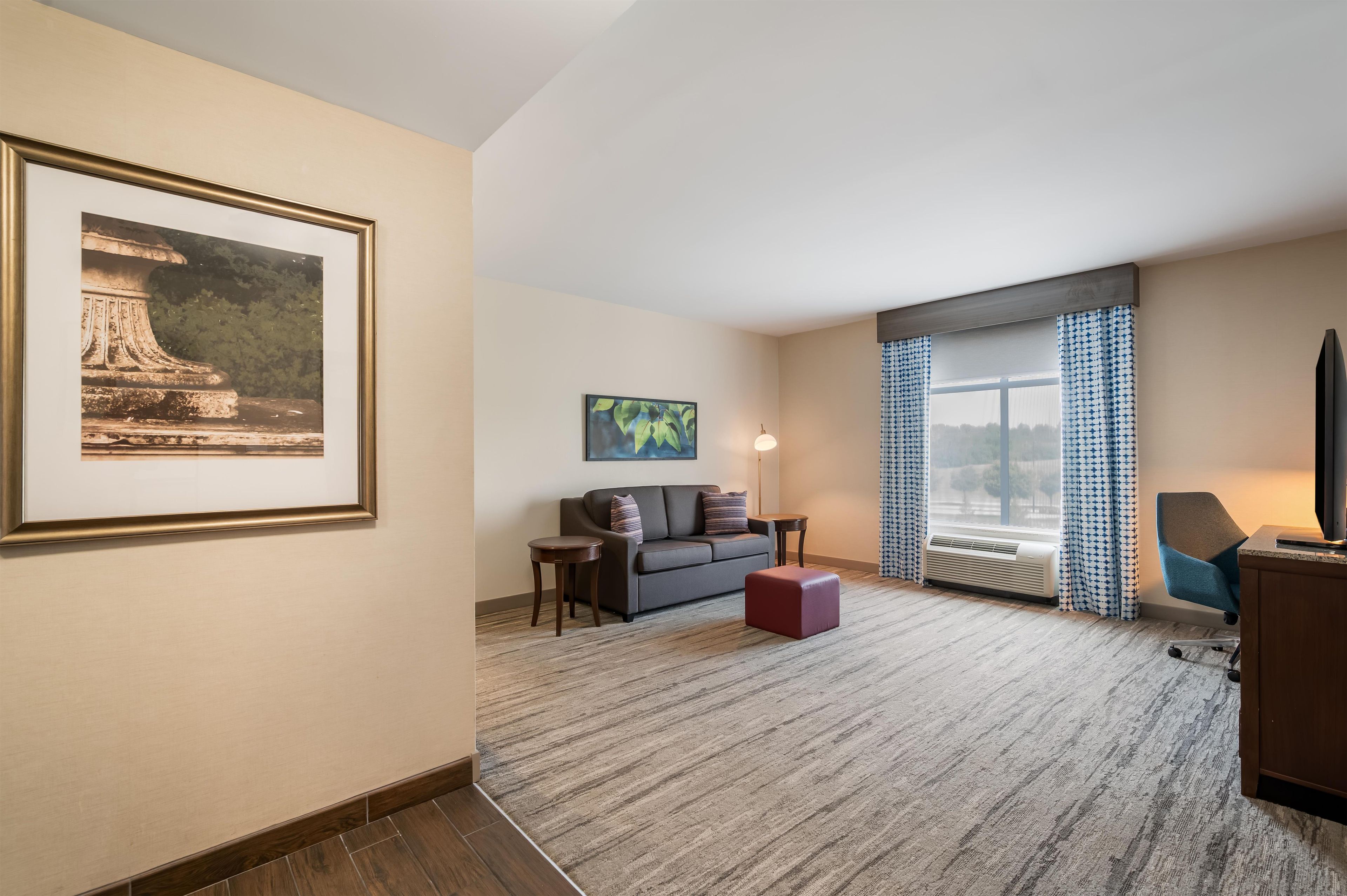 A hotel room with a large framed picture, a sofa, a small table, a lamp, and a large window with curtains.