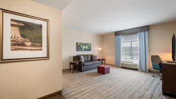 A hotel room with a large framed picture, a sofa, a small table, a lamp, and a large window with curtains.