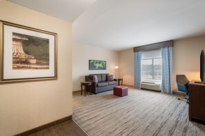 Habitación de hotel con un cuadro grande enmarcado, un sofá, una mesita, una lámpara y un ventanal con cortinas.
