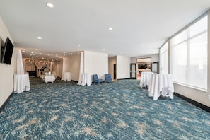 Amplia habitación con alfombra estampada, mesas con manteles blancos, una silla azul y una cocina visible a través de un panel de vidrio.