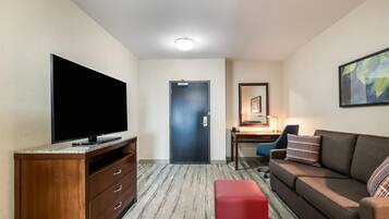 A hotel room with a flat-screen TV, a wooden dresser, a desk with a chair, a sofa, and a red ottoman.