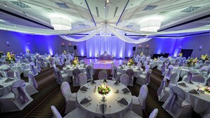 Un salón de banquetes con mesas redondas, manteles blancos y sillas adornadas con cintas moradas.