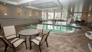 Un área de piscina interior con sillones reclinables y mesas, un letrero que prohíbe el clavado, y vistas al exterior a través de grandes ventanales.