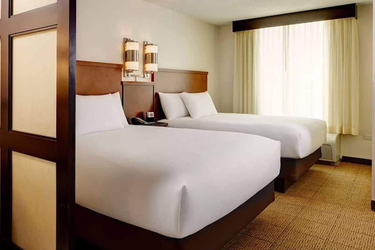 A hotel room with two beds, a headboard, bedside lamps, a nightstand, and a window with curtains.