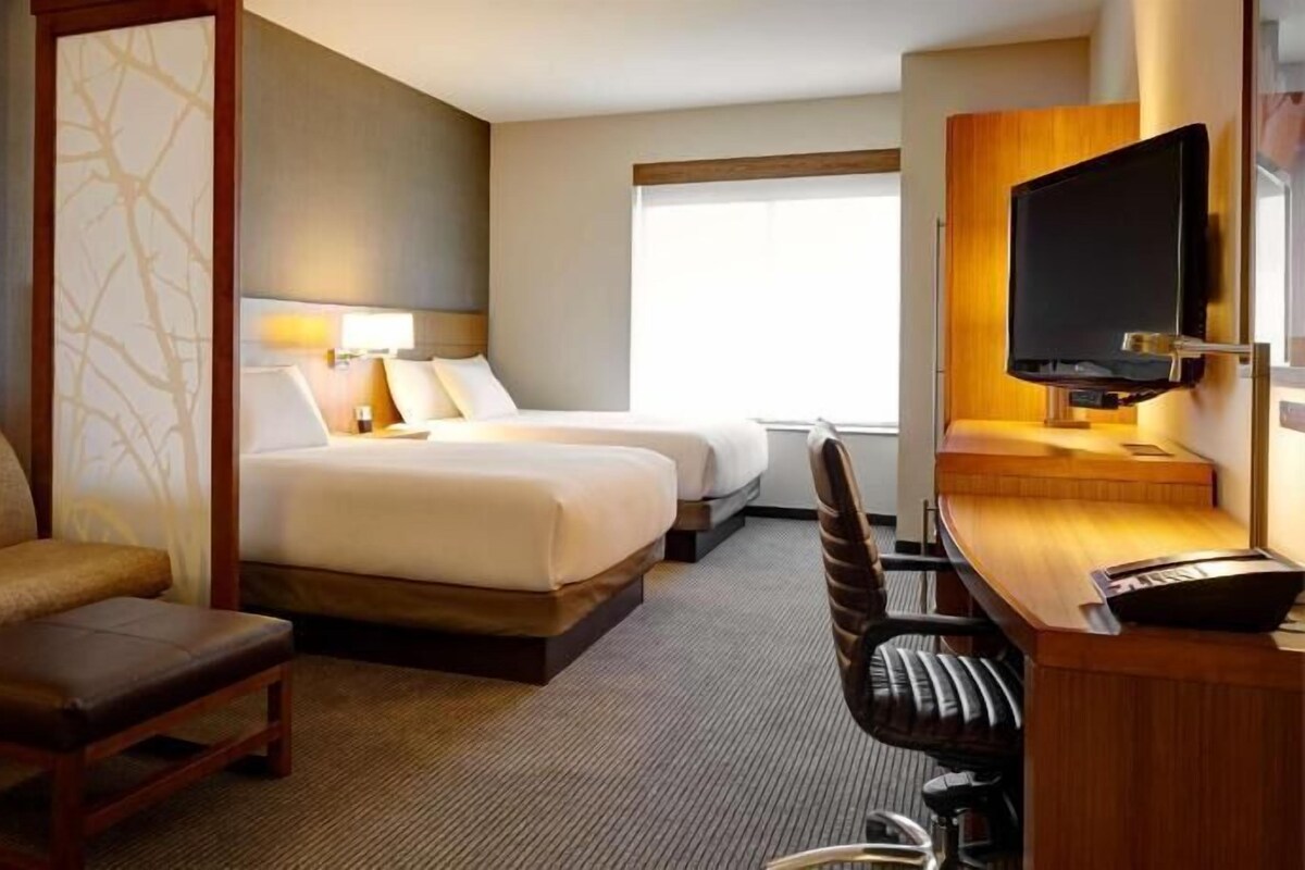 A hotel room with a sofa, a desk with a chair, two beds, a TV, and a window.