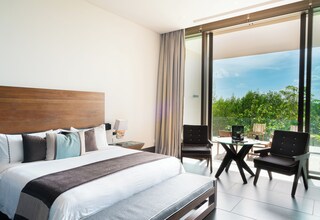 A modern hotel room with a large bed, a desk, two chairs, and a balcony with a view of greenery.