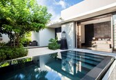 A modern outdoor pool area with a glass barrier, a seating area with a sofa and table, and a view into a well-lit interior space.
