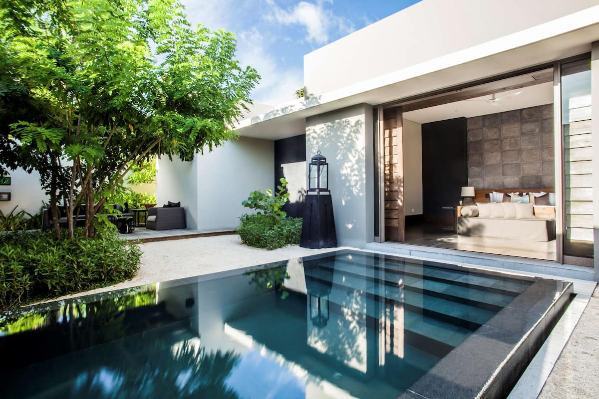 A modern outdoor pool area with a glass barrier, a seating area with a sofa and table, and a view into a well-lit interior space.