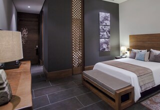A modern hotel room with a large bed, bedside tables, a bench, a decorative wall panel, and a view of the hallway.