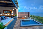 A modern outdoor pool area with a wooden deck, a seating area with cushions, and a view of the ocean.