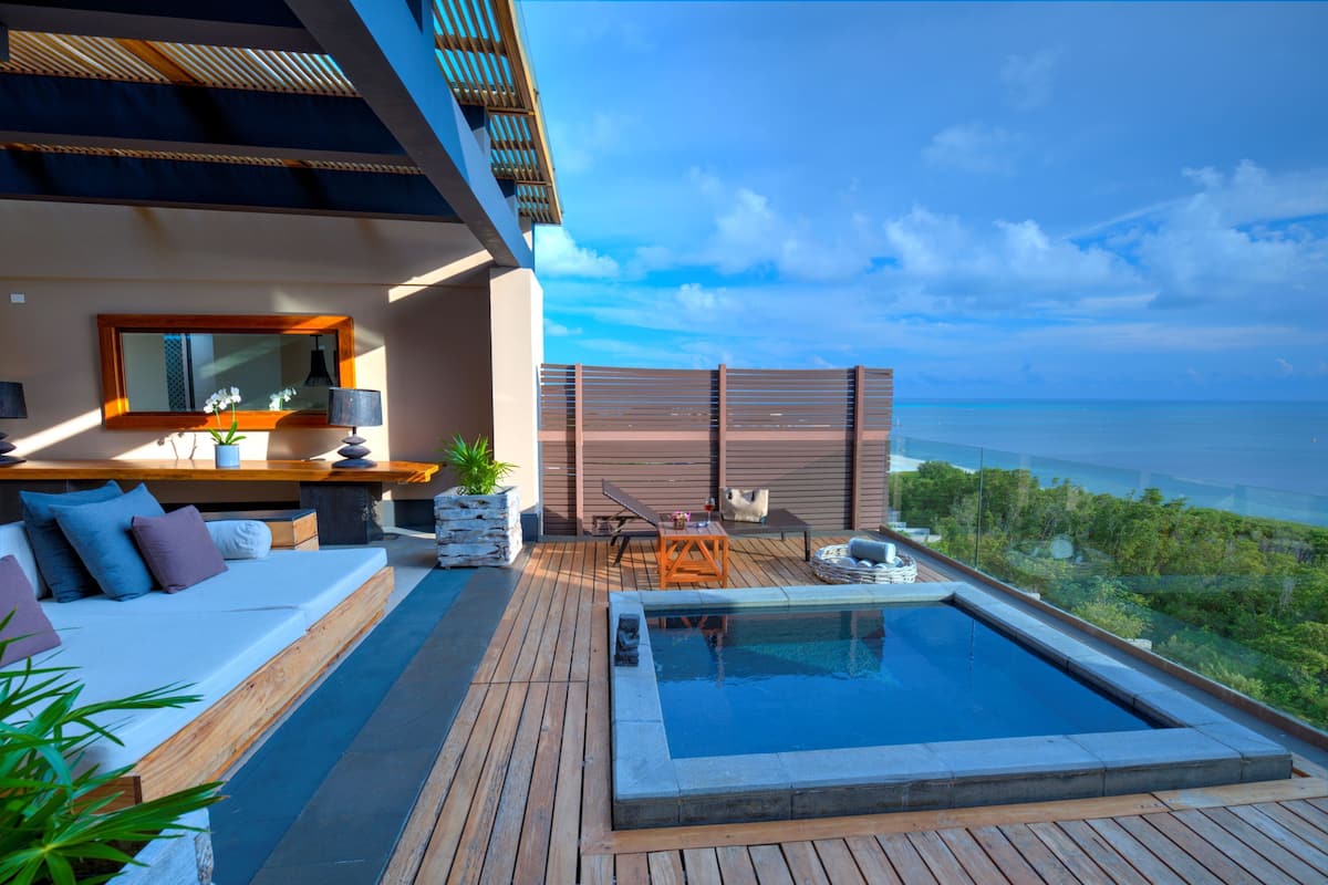 A modern outdoor pool area with a wooden deck, a seating area with cushions, and a view of the ocean.