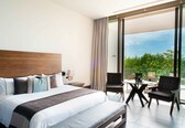 A modern hotel room with a large bed, a desk, and a balcony with a view of greenery.