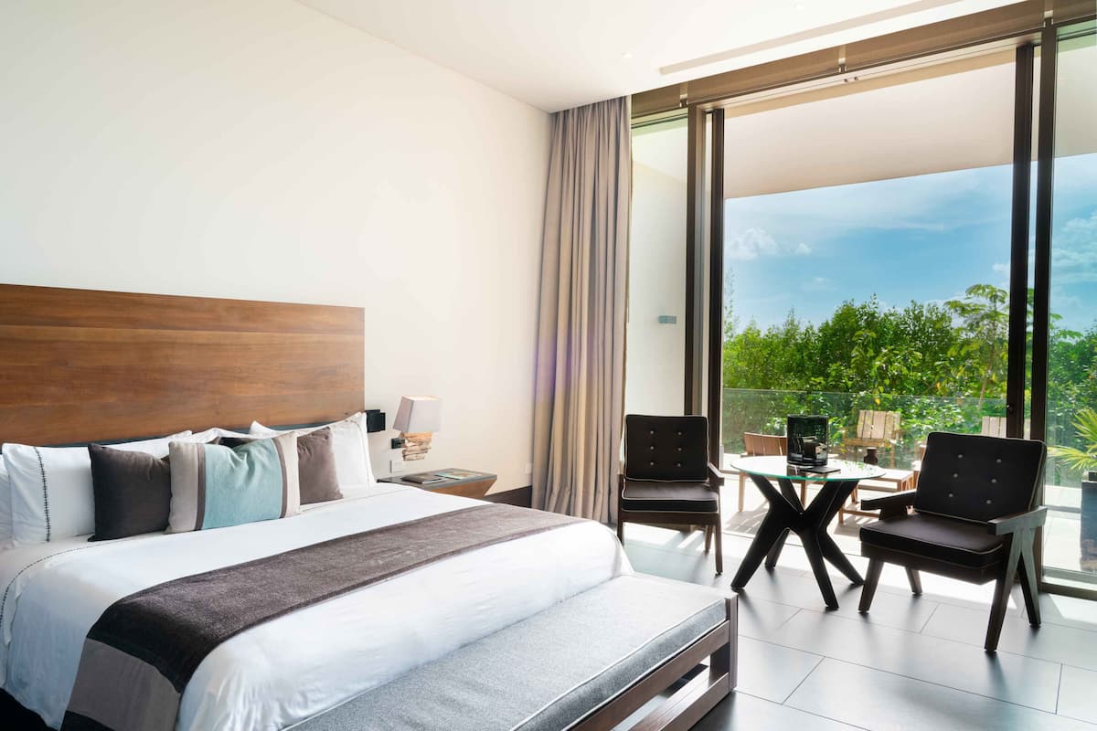 A modern hotel room with a large bed, a desk, and a balcony with a view of greenery.