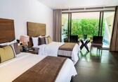 A hotel room with two beds, a desk with a phone, and a view of greenery through large windows.