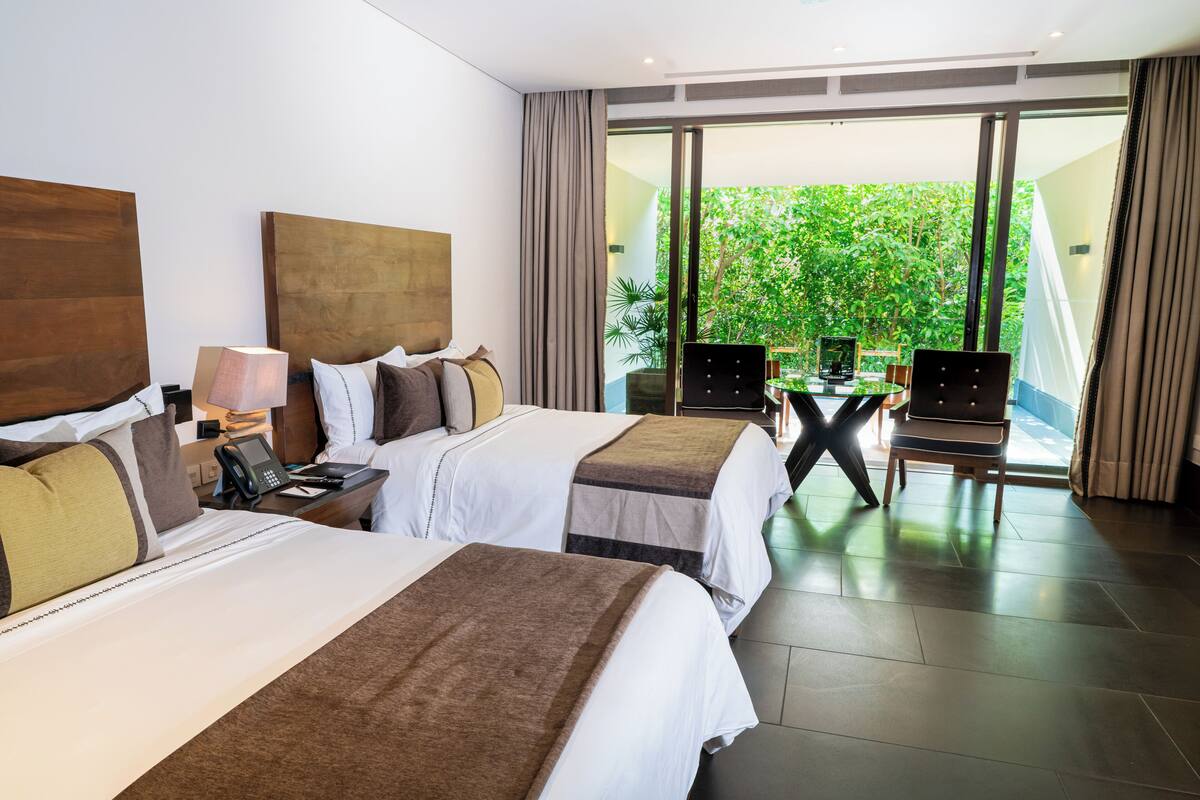 A hotel room with two beds, a desk with a phone, and a view of greenery through large windows.