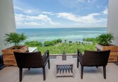 A balcony with wicker chairs and a small table, overlooking a clear blue sea and lush greenery.