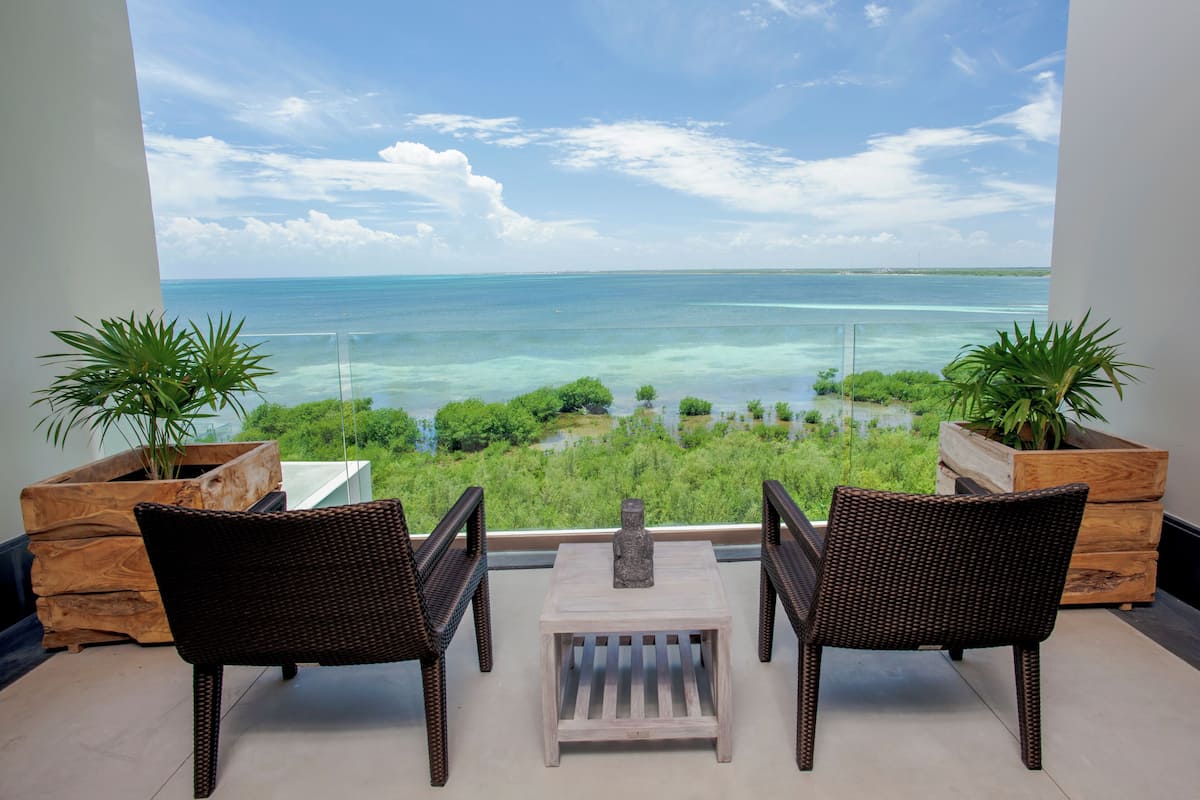 A balcony with wicker chairs and a small table, overlooking a clear blue sea and lush greenery.