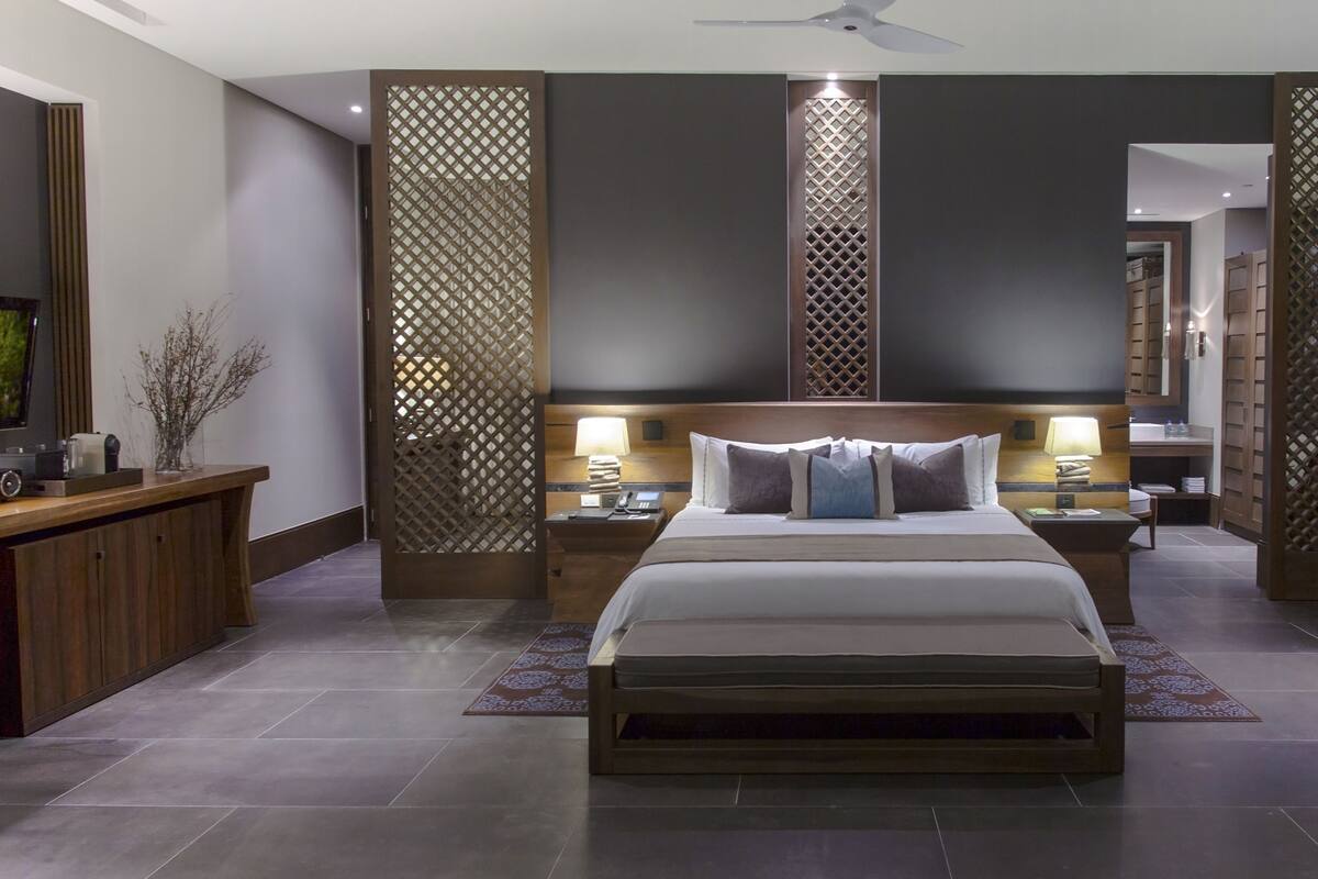 A modern hotel room with a large bed, wooden headboard, two bedside tables with lamps, a small desk, and a chair.