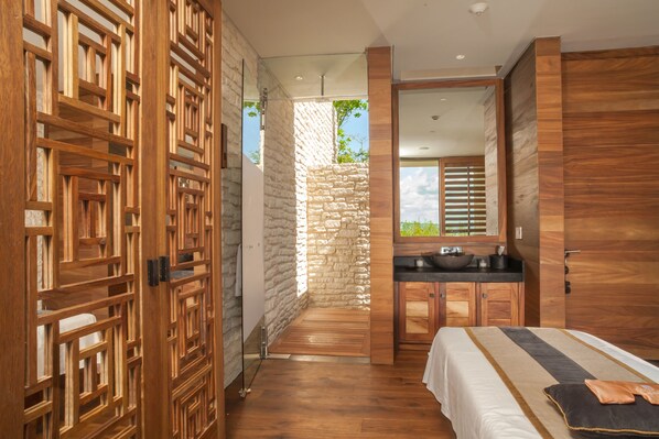 Una habitación de hotel moderna con suelo de madera, pared de acento de piedra, una cama grande, un armario de madera y un baño con tocador y lavamanos de madera.