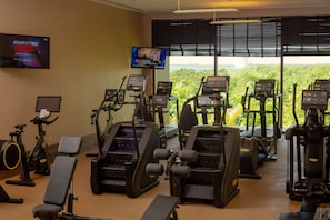 A gym with various exercise equipment, a television displaying a sports event, and a view of greenery outside.