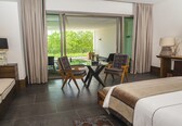 A hotel room with a bed, a sofa, a dining table with chairs, a lamp, and a view of greenery through the glass door.