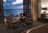 A hotel room with a bed, two armchairs, a small table, a lamp, and a view of the sea.
