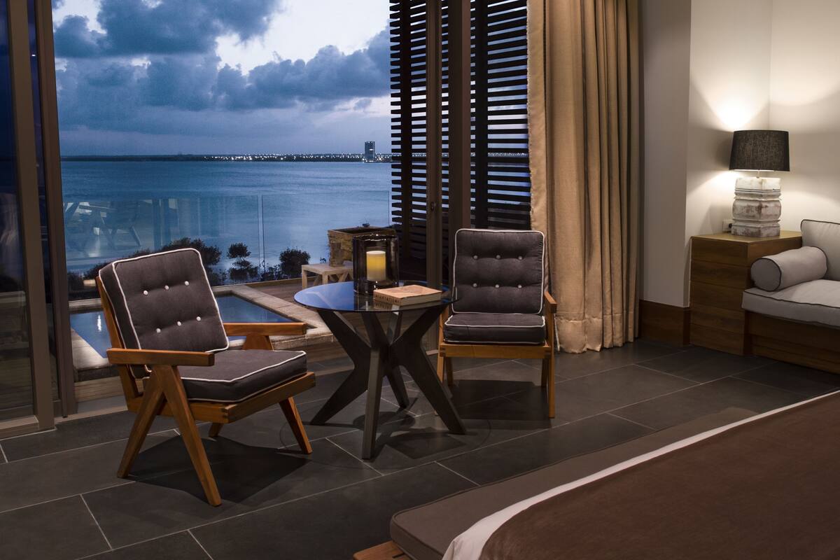 A hotel room with a bed, two armchairs, a small table, a lamp, and a view of the sea.