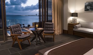 A hotel room with a bed, two armchairs, a small table, a lamp, and a view of the sea.