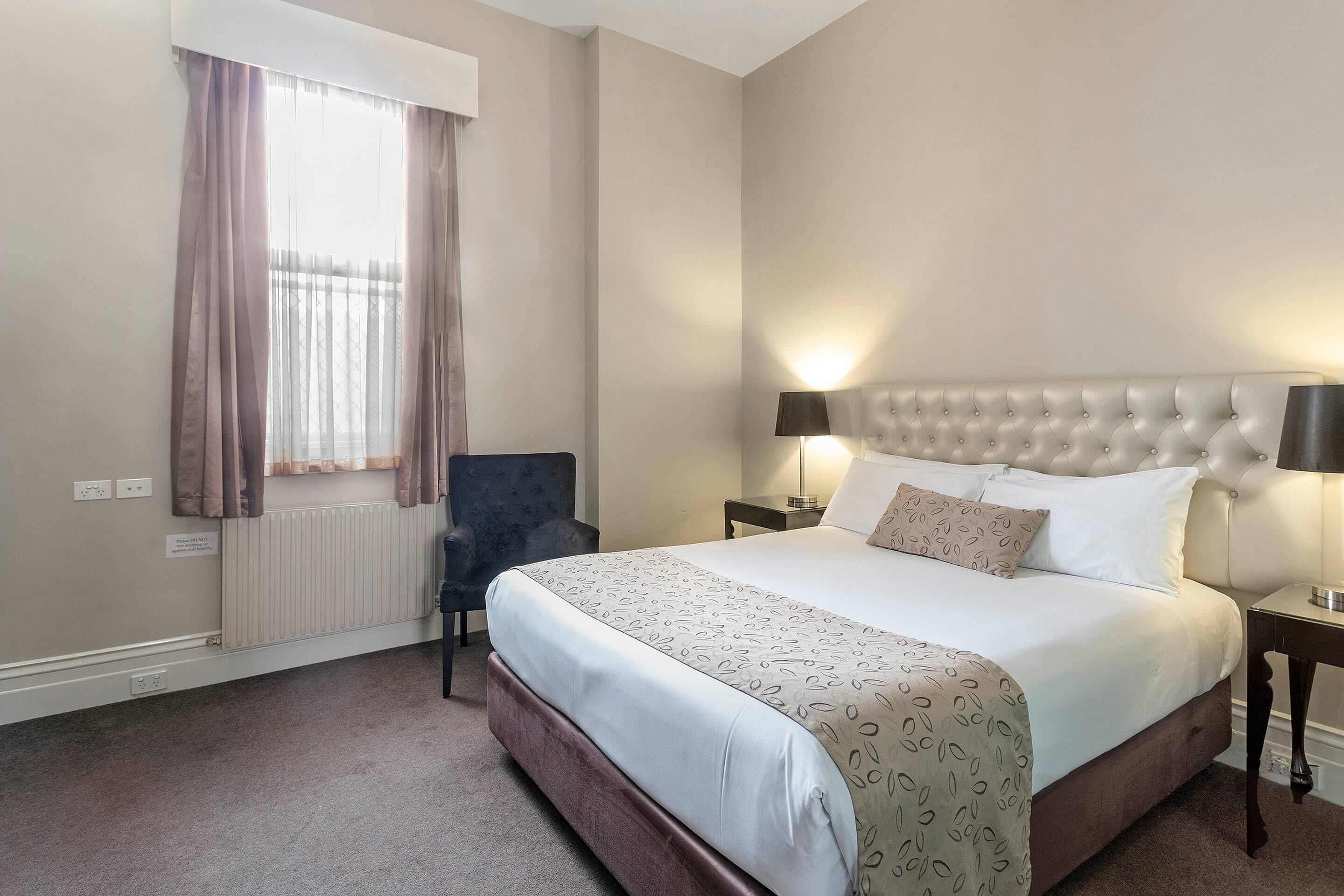 Una habitación de hotel con cama, una silla, mesita de noche, lámpara y ventana con cortinas.