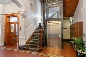 A hotel lobby with a wooden staircase, an elevator, a reception desk, and a potted plant.