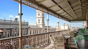 A covered terrace with wicker furniture, ornate railings, and a view of historic buildings.