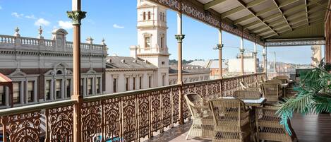 A covered terrace with wicker furniture, ornate railings, and a view of historic buildings.