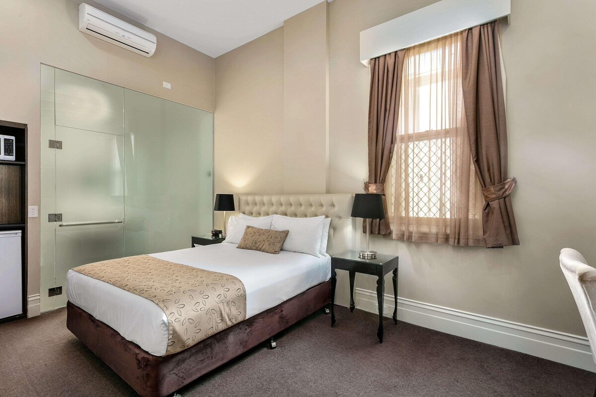 A hotel room with a bed, bedside table, lamp, and a window with curtains.