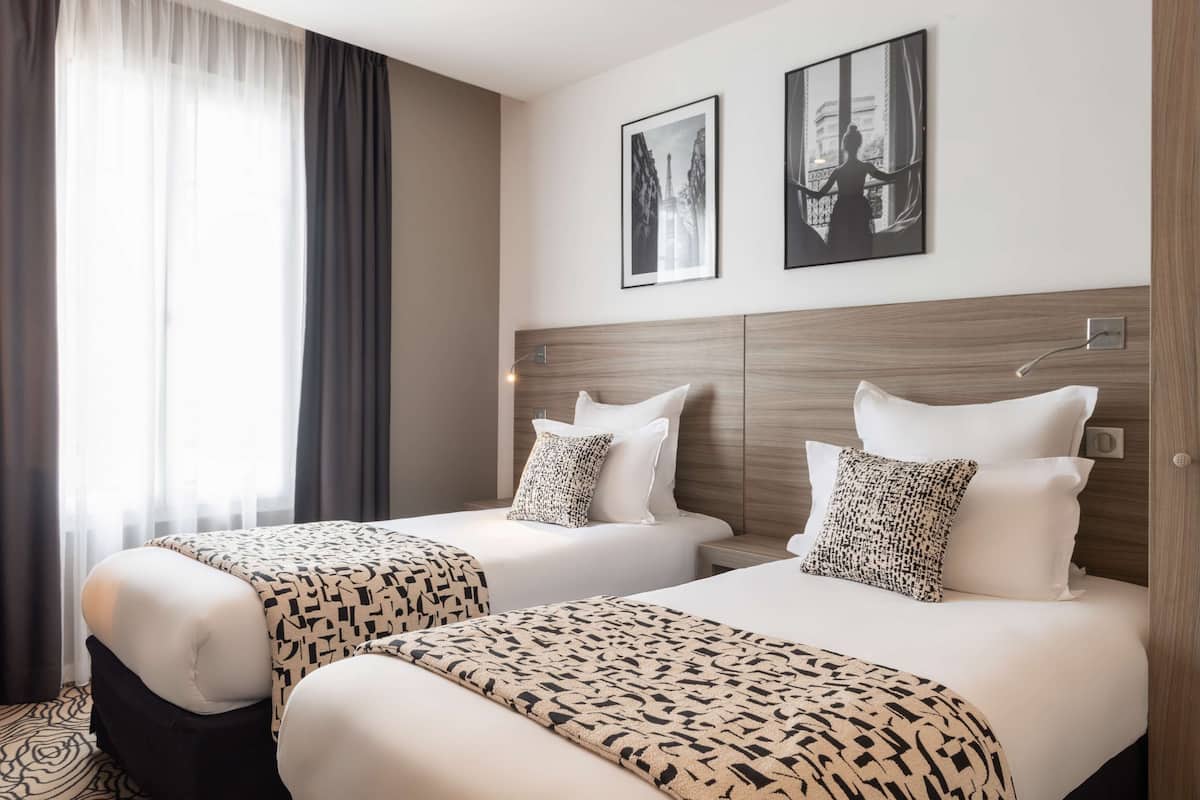Une chambre d’hôtel avec deux lits, chacun agrémenté d’une couvre-lit à motifs et de coussins assortis. Deux tableaux encadrés ornent le mur, il y a une fenêtre avec des rideaux et une tête de lit en bois.