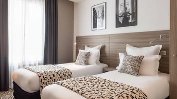 Une chambre d’hôtel avec deux lits, chacun agrémenté d’une couvre-lit à motifs et de coussins assortis. Deux tableaux encadrés ornent le mur, il y a une fenêtre avec des rideaux et une tête de lit en bois.